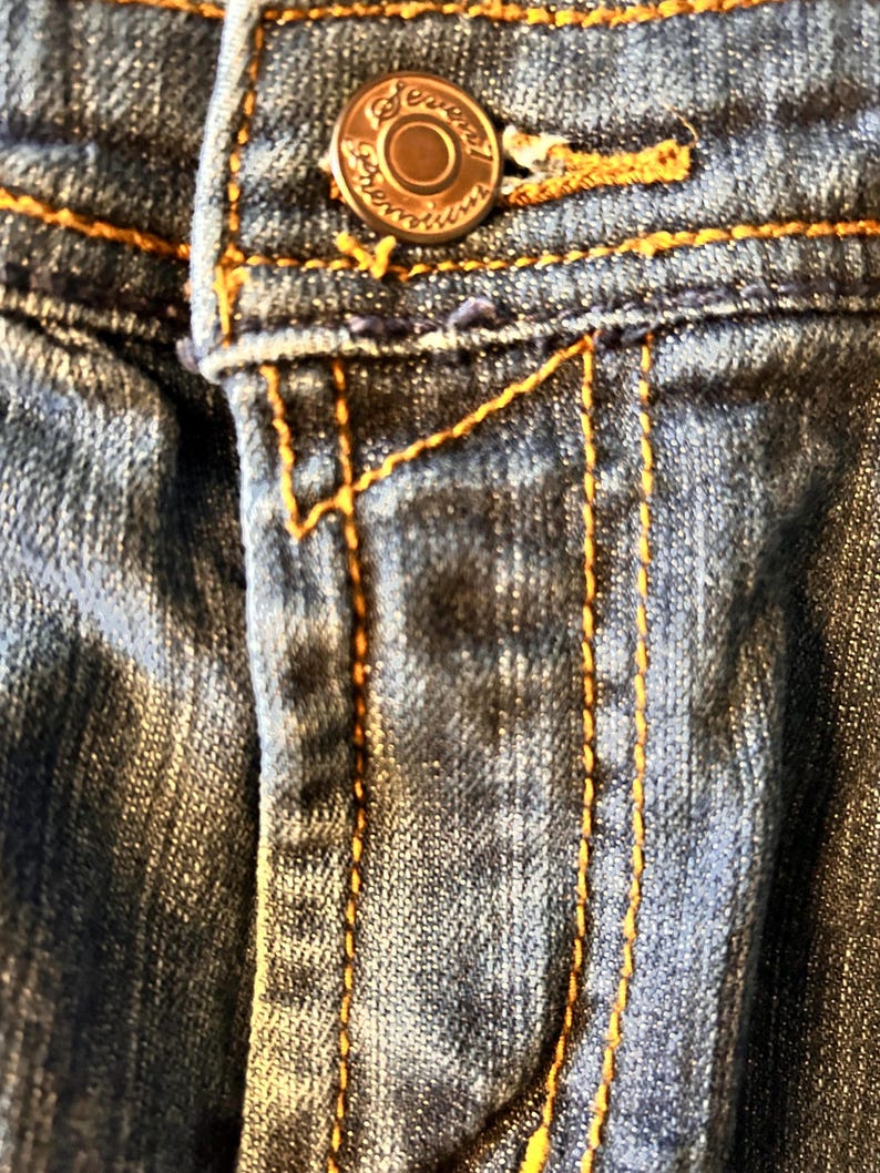 May include: Close-up of denim jeans with a button and orange stitching. The button is a copper color with the word "Seven" around the edge. The jeans have a classic five-pocket design.