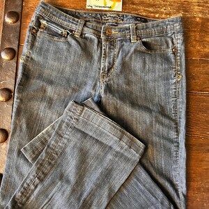 May include: Blue denim jeans with a classic five-pocket design. The jeans have a button and zipper closure, belt loops, and decorative stitching. The jeans are folded on a wooden surface. A vintage sticker is in the background.