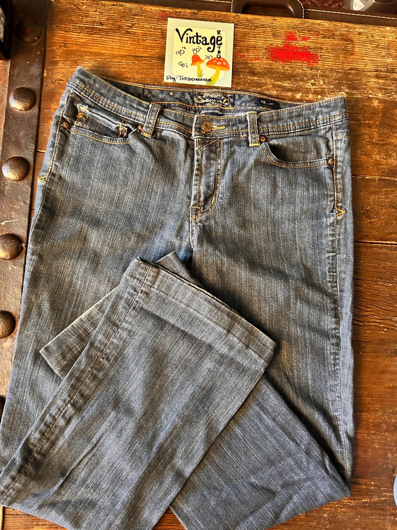 May include: Blue denim jeans with a classic five-pocket design. The jeans have a button and zipper closure, belt loops, and decorative stitching. The jeans are folded on a wooden surface. A vintage sticker is in the background.