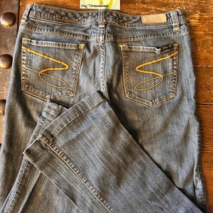 May include: Dark wash denim jeans with gold stitching and a flared leg. The back pockets feature a distinctive gold 'S' design. A small tag is visible on the back waistband. The jeans are laid out on a wooden surface. A vintage sticker is in the background.