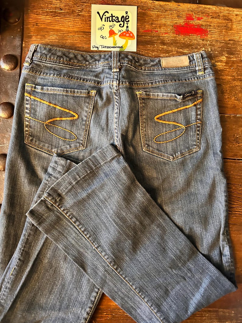 May include: Dark wash denim jeans with gold stitching and a flared leg. The back pockets feature a distinctive gold 'S' design. A small tag is visible on the back waistband. The jeans are laid out on a wooden surface. A vintage sticker is in the background.