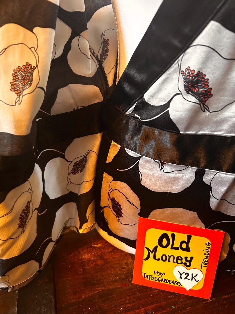 May include: Black and white floral print fabric with a satin finish. The design features large white flowers with orange and red centers against a black background. A tag with the words "Old Money" and "Y2K" is visible.