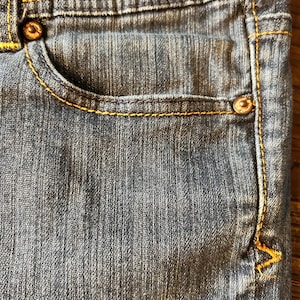 May include: Close-up of a section of blue denim jeans. The jeans feature a small pocket, metal rivets, and yellow stitching. The fabric has a textured appearance.