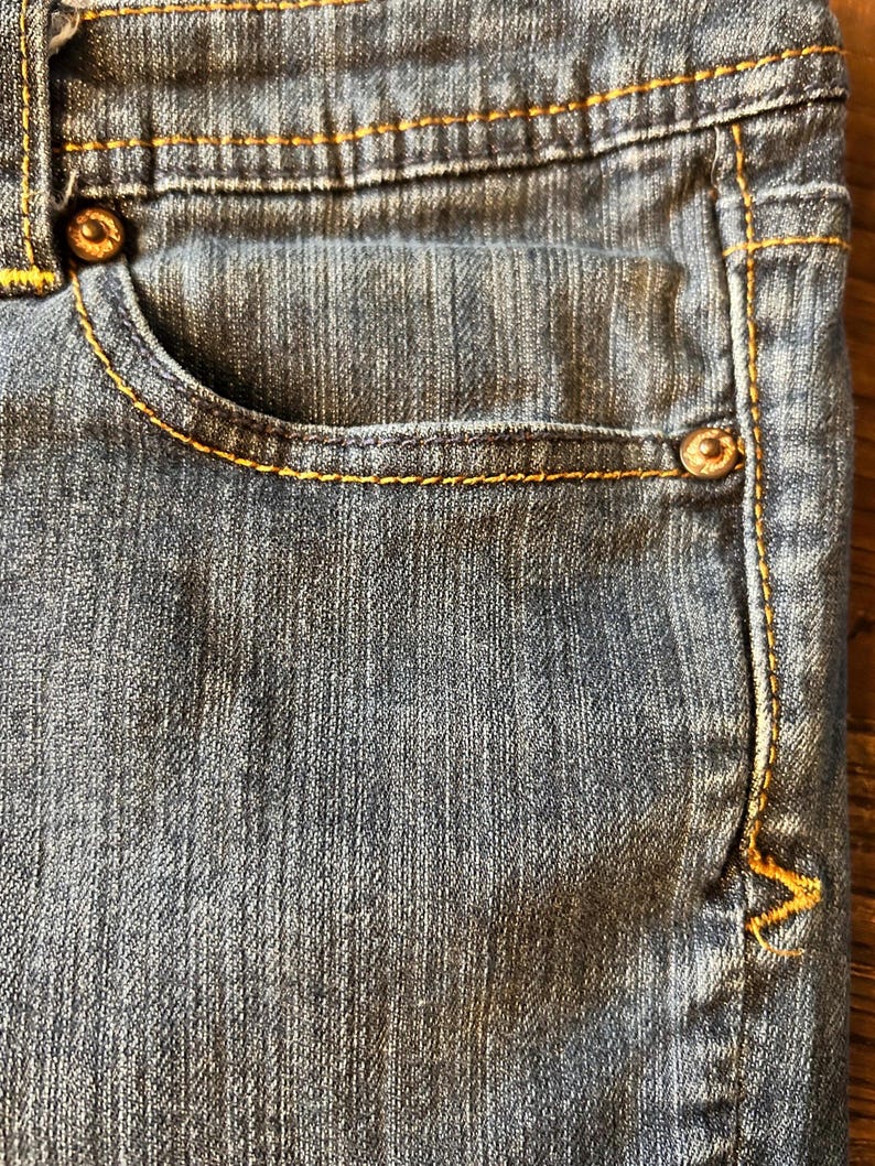 May include: Close-up of a section of blue denim jeans. The jeans feature a small pocket, metal rivets, and yellow stitching. The fabric has a textured appearance.