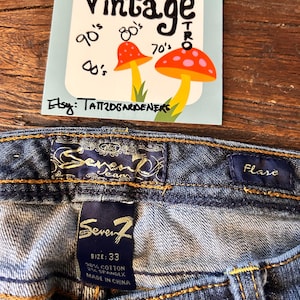 May include: Close-up of blue denim jeans with a flare cut. The jeans have a label that says "Seven7" and a size of 33. A vintage-style sticker with mushrooms and the word "Vintage" is also visible.