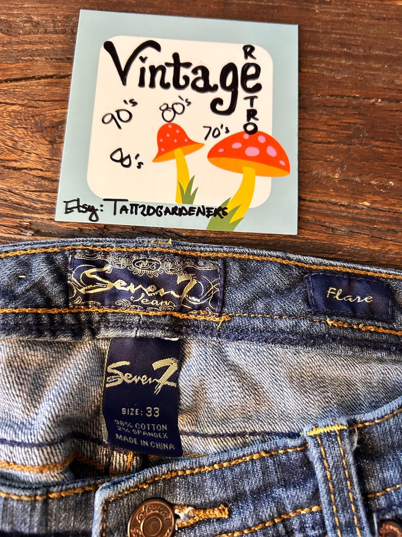May include: Close-up of blue denim jeans with a flare cut. The jeans have a label that says "Seven7" and a size of 33. A vintage-style sticker with mushrooms and the word "Vintage" is also visible.
