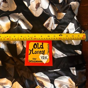 May include: Black satin wrap top with a white floral pattern and a black trim. The top features a bold floral design with brown centers. A yellow and red sticker with the text "Old Money" and "Y2K" is visible.