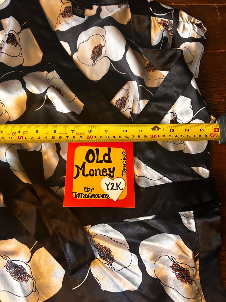May include: Black satin wrap top with a white floral pattern and a black trim. The top features a bold floral design with brown centers. A yellow and red sticker with the text "Old Money" and "Y2K" is visible.