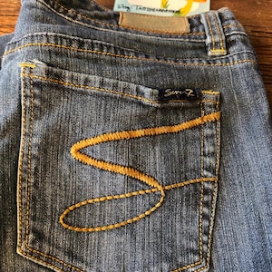 May include: Close-up of blue denim jeans with a pocket featuring a decorative gold-colored stitch design. The jeans have a small, dark blue label with the brand name "Seven7". A vintage tag with mushroom illustrations is visible.