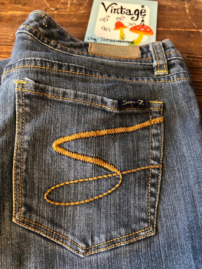 May include: Close-up of blue denim jeans with a pocket featuring a decorative gold-colored stitch design. The jeans have a small, dark blue label with the brand name "Seven7". A vintage tag with mushroom illustrations is visible.