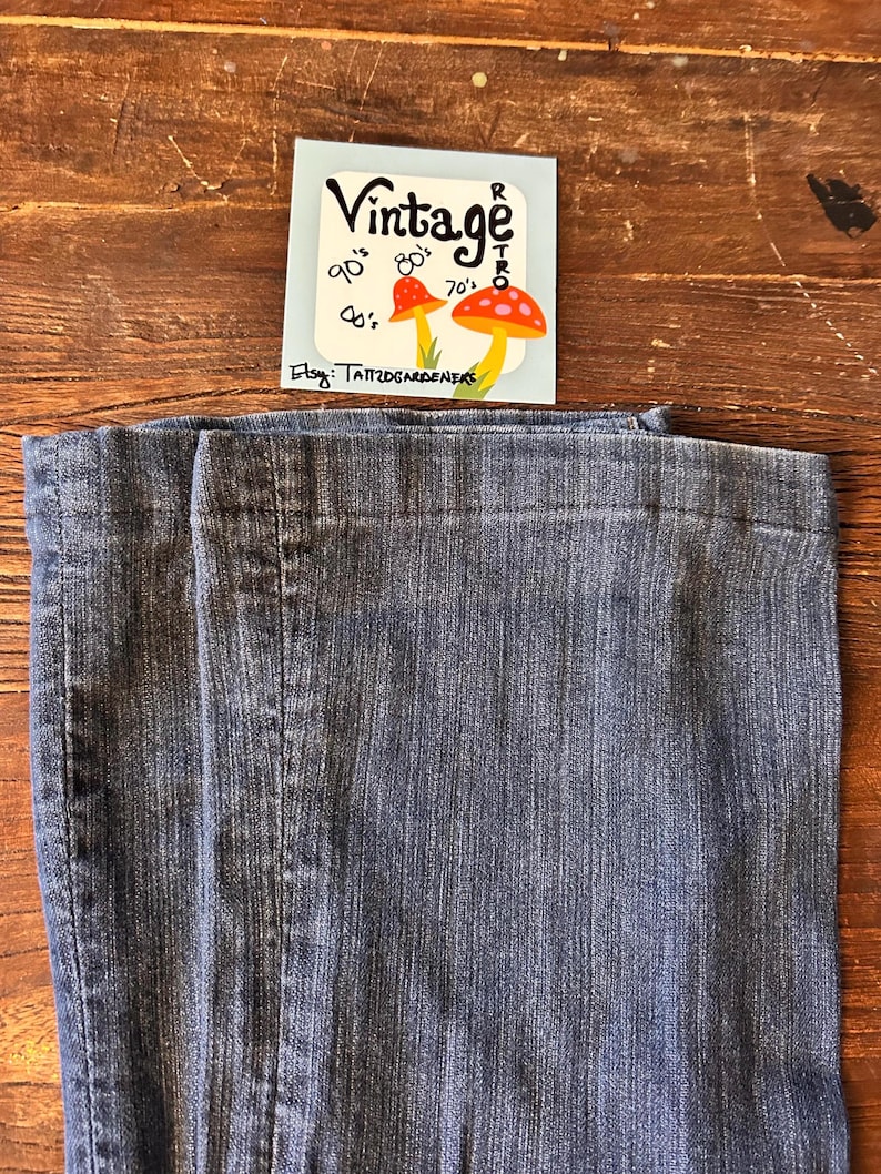 May include: Close-up of blue denim jeans. A small tag with the word "Vintage" and mushroom illustrations is visible. The tag also includes the text "90's, 80's, 70's, 60's" and "Etsy: TATTOOGARDENERS".