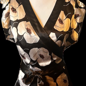 May include: Black and white floral wrap top with a V-neckline and short sleeves. The top features a satin bow at the waist and a contrasting black trim. The floral pattern includes white and gold flowers with brown centers.
