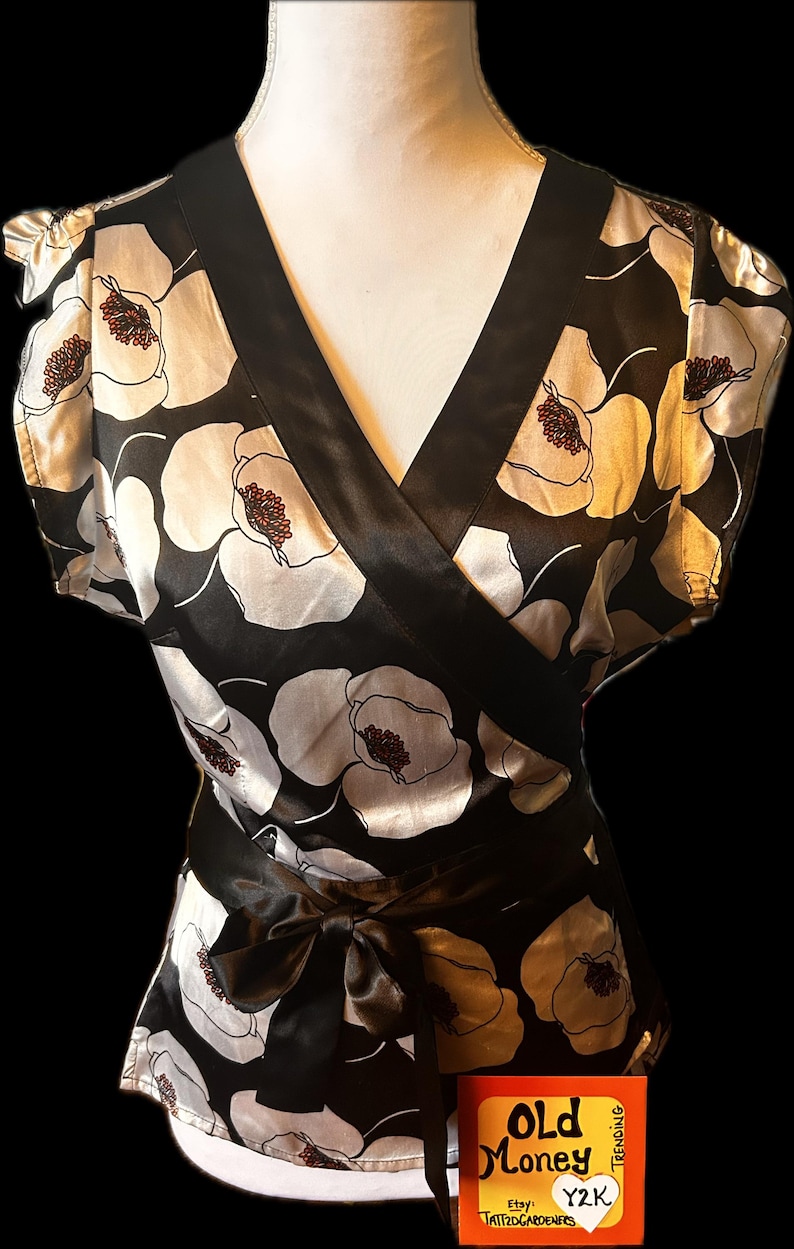 May include: Black and white floral wrap top with a V-neckline and short sleeves. The top features a satin bow at the waist and a contrasting black trim. The floral pattern includes white and gold flowers with brown centers.