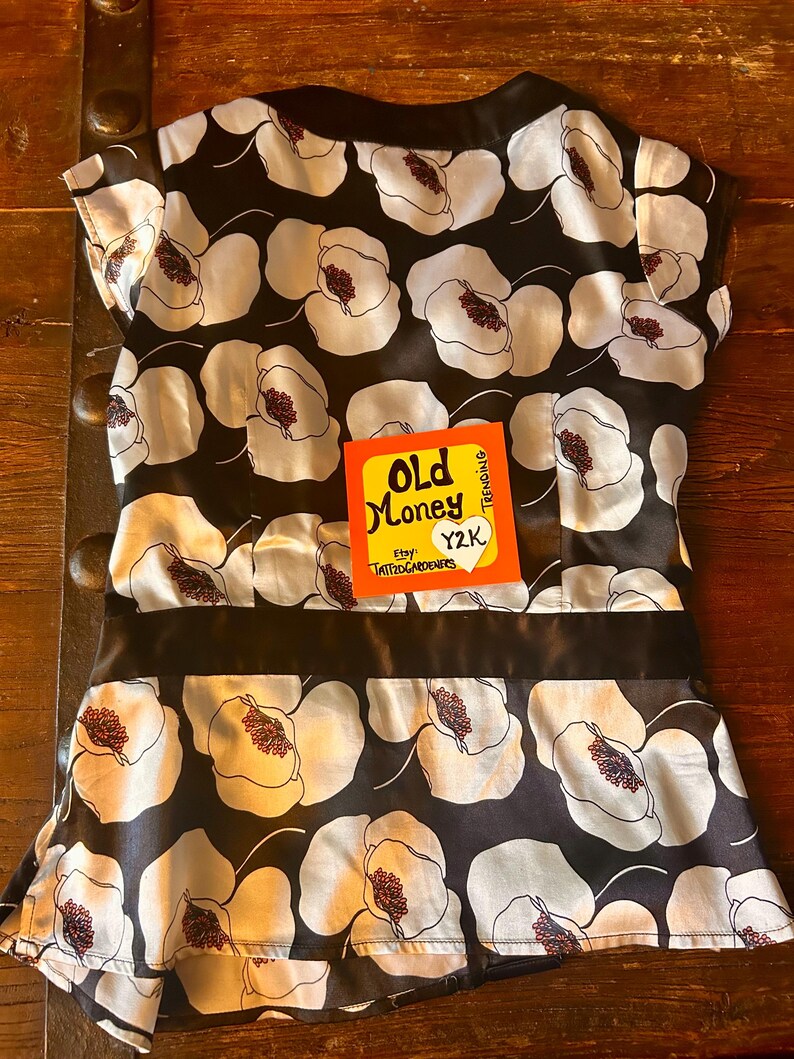 May include: A black and white floral print top with a black satin band. The top features a pattern of large white flowers with red centers on a black background. The tag reads "Old Money" and "Y2K".