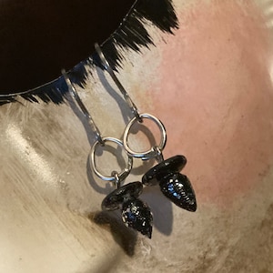 May include: A pair of dangle earrings featuring silver-tone hoops and black, glitter-covered acorn-shaped charms. The earrings are displayed against a neutral background, highlighting their design and sparkle.
