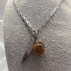 Puede incluir: Un collar plateado con una cadena de eslabones rectangulares. El colgante presenta un pequeño cuchillo y una piedra preciosa marrón en forma de lágrima. El collar se muestra sobre una superficie gris.