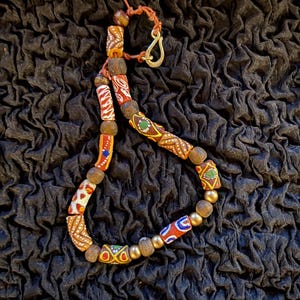 Puede incluir: Un collar artesanal con una variedad de cuentas coloridas y estampadas. El collar incluye cuentas cilíndricas y redondas en tonos rojos, naranjas, amarillos y verdes, ensartadas con un cierre y un cordón dorados.