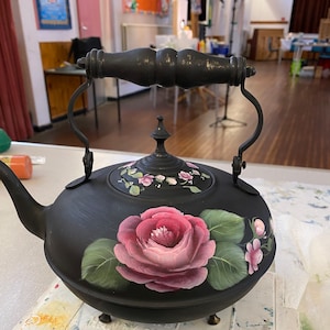 May include: Black teapot with a painted design of pink roses and green leaves. The teapot has a black handle and a black lid.