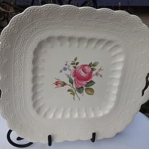 May include: A white, square serving platter with a scalloped edge and a floral rose design. The platter has a decorative, embossed border and two curved handles. The center features a pink rose with green leaves and small blue flowers.