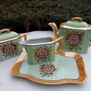 May include: A vintage tea set featuring a teapot, sugar bowl, creamer, and serving tray. The set is light green with gold trim and a floral design in pink, yellow, and green. The teapot measures approximately 6 inches in height.