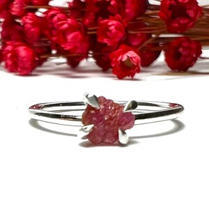 May include: A silver ring featuring a raw, uncut ruby gemstone. The ruby is a deep pinkish-red color and is held in place by four silver prongs. The ring is set against a white background with red flowers.