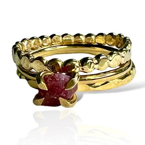 Może przedstawiać: Zestaw trzech złotych pierścionków z różowym kamieniem szlachetnym w środku. Pierścionki są ułożone jeden na drugim i mają fakturowany wzór.