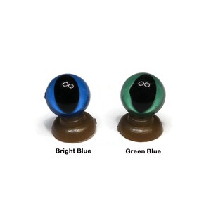May include: Two plastic doll eyes on display stands. The eye on the left is bright blue with a black pupil. The eye on the right is green blue with a black pupil.