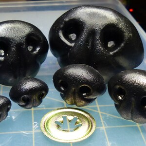 May include: A set of six black plastic animal noses, varying in size. The noses have two nostrils and a raised ridge in the center. The noses are commonly used for craft projects and toy making.