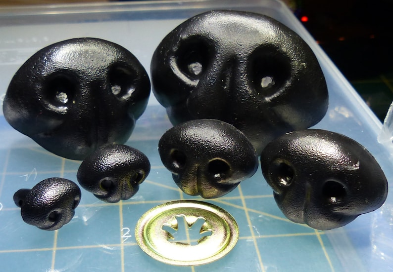 May include: A set of six black plastic animal noses, varying in size. The noses have two nostrils and a raised ridge in the center. The noses are commonly used for craft projects and toy making.