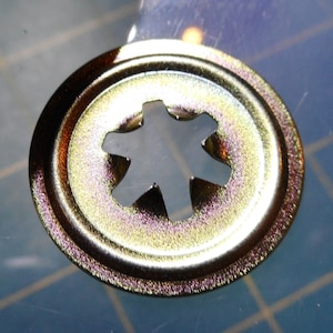 May include: A gold-colored metal fastener with a star-shaped hole in the center. The fastener has a raised edge around the perimeter.