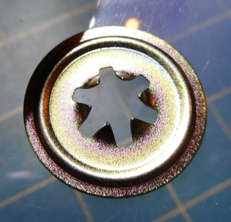 May include: A gold-colored metal fastener with a star-shaped hole in the center. The fastener has a raised edge around the perimeter.
