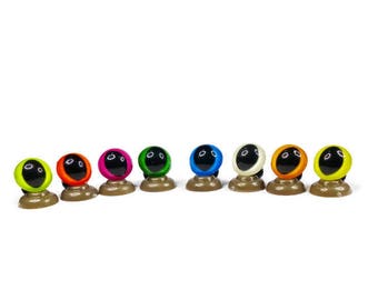 May include: Ten colorful plastic safety eyes with black pupils, mounted on brown plastic bases. The eyes are arranged in a row from left to right, with the colors being neon green, orange, pink, green, blue, white, orange, yellow, and black.