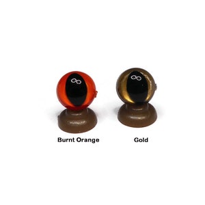 May include: Two plastic doll eyes on display stands. The eye on the left is burnt orange with a black pupil. The eye on the right is gold with a black pupil.