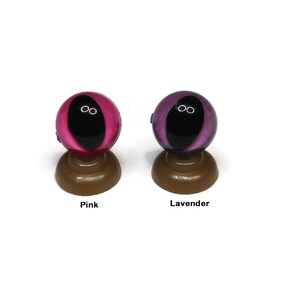 May include: Two glass eyes on display stands. The eye on the left is pink with a black pupil. The eye on the right is lavender with a black pupil.