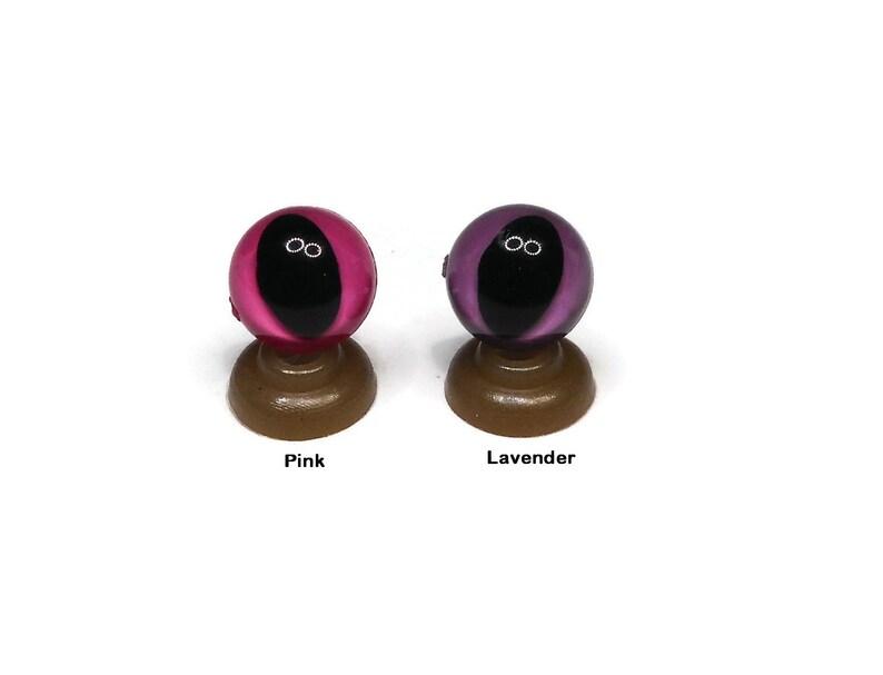 May include: Two glass eyes on display stands. The eye on the left is pink with a black pupil. The eye on the right is lavender with a black pupil.