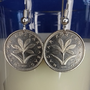 May include: Silver earrings featuring a coin with a floral design and the text "KOZTARSASAG" and "MAGYAR" on either side. The coin is dated "1997" on the left earring and "2000" on the right earring.