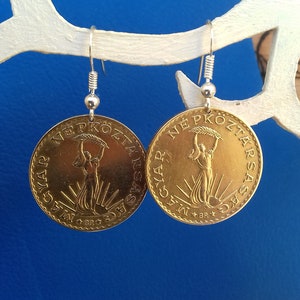 May include: A pair of gold-colored earrings featuring a round coin design with the text "NE PKOZTAR SAS" and "GYAR  AM  DBD" on each earring. The earrings are hanging from silver hooks.