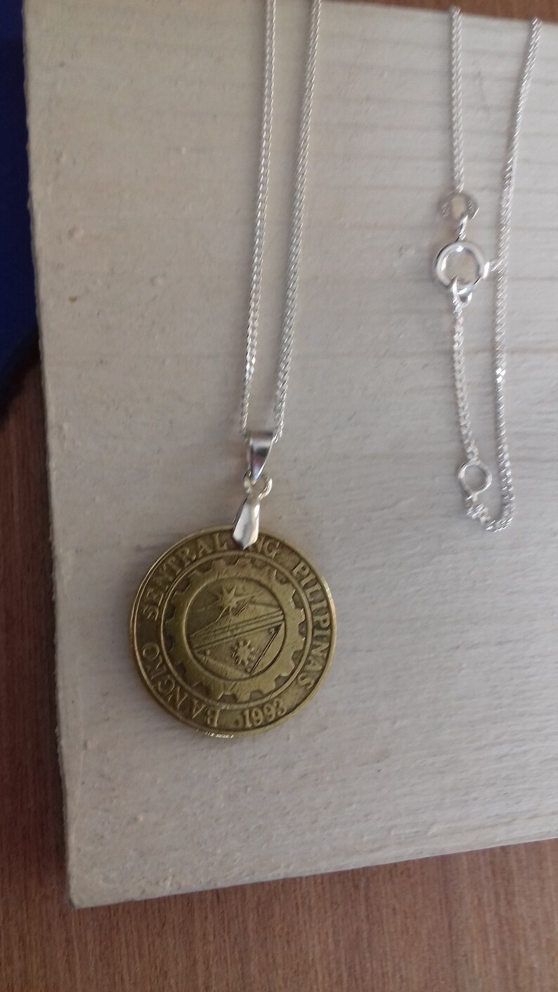 May include: A silver chain necklace with a round pendant featuring the seal of the Central Bank of the Philippines. The seal is gold in colour and features a gear with a sun and a star inside. The text "Bangko Sentral ng Pilipinas" and "1993" are visible on the pendant.