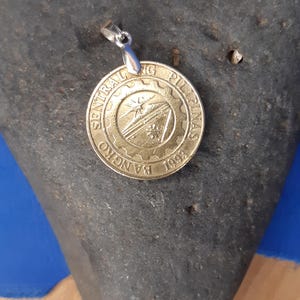 May include: A gold-coloured pendant featuring the logo of the Bangko Sentral ng Pilipinas, a central bank in the Philippines. The logo depicts a stylised mountain range with a sun rising behind it. The pendant is attached to a silver chain.
