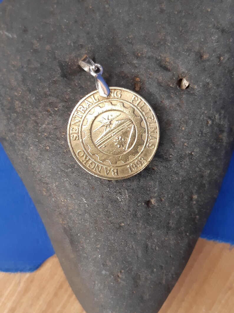 May include: A gold-coloured pendant featuring the logo of the Bangko Sentral ng Pilipinas, a central bank in the Philippines. The logo depicts a stylised mountain range with a sun rising behind it. The pendant is attached to a silver chain.