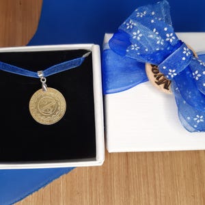 May include: A gold pendant necklace with a round, detailed design is displayed in a black velvet box. The box is surrounded by a blue ribbon and a white gift box with a blue bow.