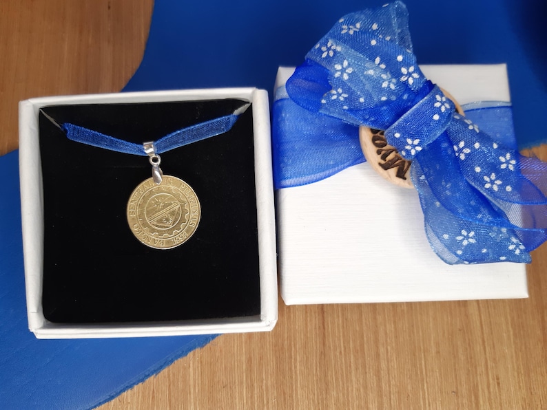 May include: A gold pendant necklace with a round, detailed design is displayed in a black velvet box. The box is surrounded by a blue ribbon and a white gift box with a blue bow.