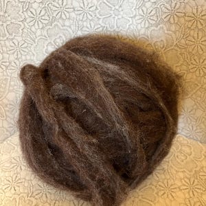 Könnte beinhalten: Ein Knäuel dunkelbrauner Wollroving, dicht gepackt und strukturiert. Die Wolle wirkt weich und flauschig, ideal für Kunsthandwerksprojekte wie Nadelfilzen oder Spinnen. Das Roving hat eine satte, erdige Farbe.