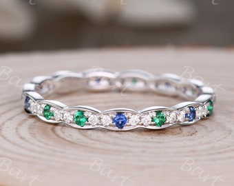 May include: A delicate eternity band featuring a row of alternating emerald green and sapphire blue gemstones set in a silver band with a scalloped edge.