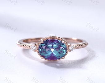 May include: A rose gold ring with an oval-shaped alexandrite gemstone surrounded by a delicate beaded band. Two smaller round diamonds are set on either side of the center stone.
