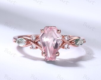 May include: A rose gold ring with a pink gemstone set in a coffin-shaped setting. The ring has a twisted band with two smaller green gemstones on each side of the main stone.