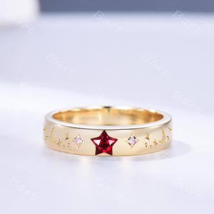 May include: A gold band ring featuring a central red star-shaped gemstone, flanked by two small iridescent round stones. The surface is engraved with celestial motifs including crescent moons and various star shapes surrounding the gemstones.