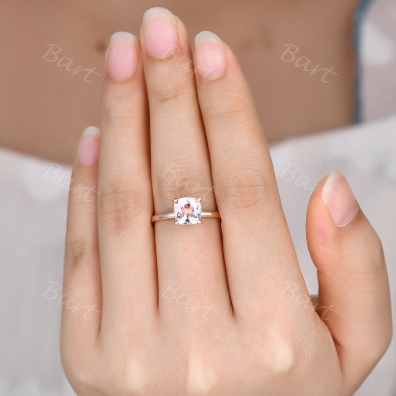 May include: A rose gold engagement ring with a square-shaped, light pink gemstone.