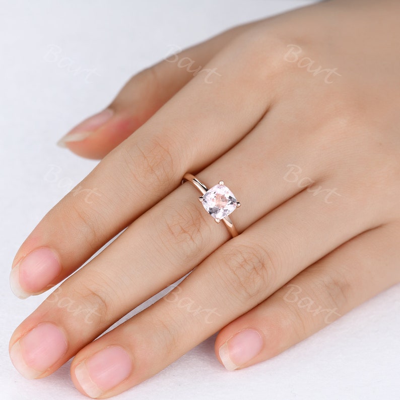 May include: A rose gold engagement ring with a square-shaped, light pink gemstone.