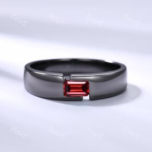 Op de afbeelding: Een zwarte ring met een rechthoekige, rode edelsteen. De ring heeft een modern, minimalistisch ontwerp met een gladde, gepolijste afwerking. De edelsteen is in het midden van de ring gezet, wat een opvallend contrast creëert.